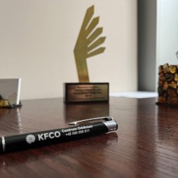 KANCELARIA FINANSOWA CENTRUM ODDŁUŻEŃ SPÓŁKA Z OGRANICZONĄ ODPOWIEDZIALNOŚCIĄ - Biuro: długopis KFCO na drewnianym biurku, w tle nagroda, wizytownik z bursztynem i pojemnik na długopisy z bursztynu. Stonowana kolorystyka, blik światła na blacie.