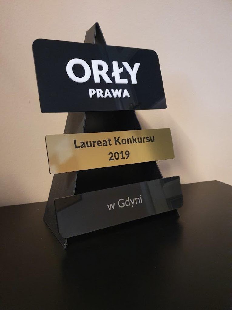 Statuetka 'Orły Prawa' dla Laureata Konkursu 2019 w Gdyni, wykonana z czarnego i złotego akrylu, stojąca na ciemnym blacie.