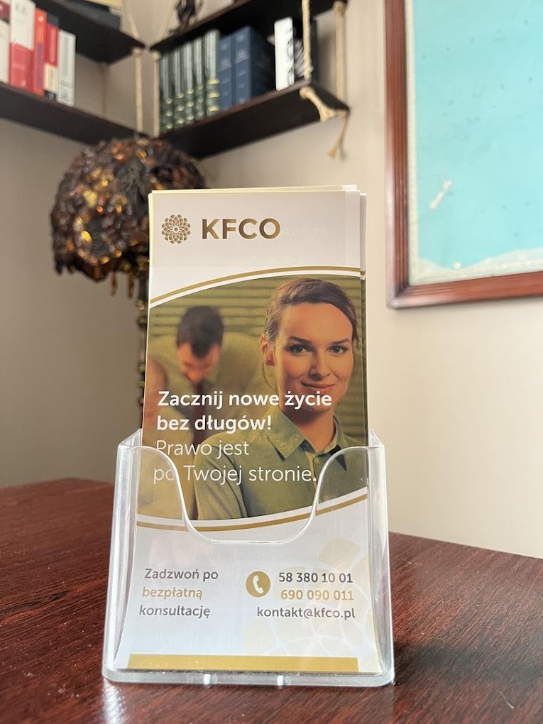 Ulotka KFCO w przezroczystym stojaku na biurku, z hasłem 'Zacznij nowe życie bez długów! Prawo jest po Twojej stronie.' i danymi kontaktowymi.