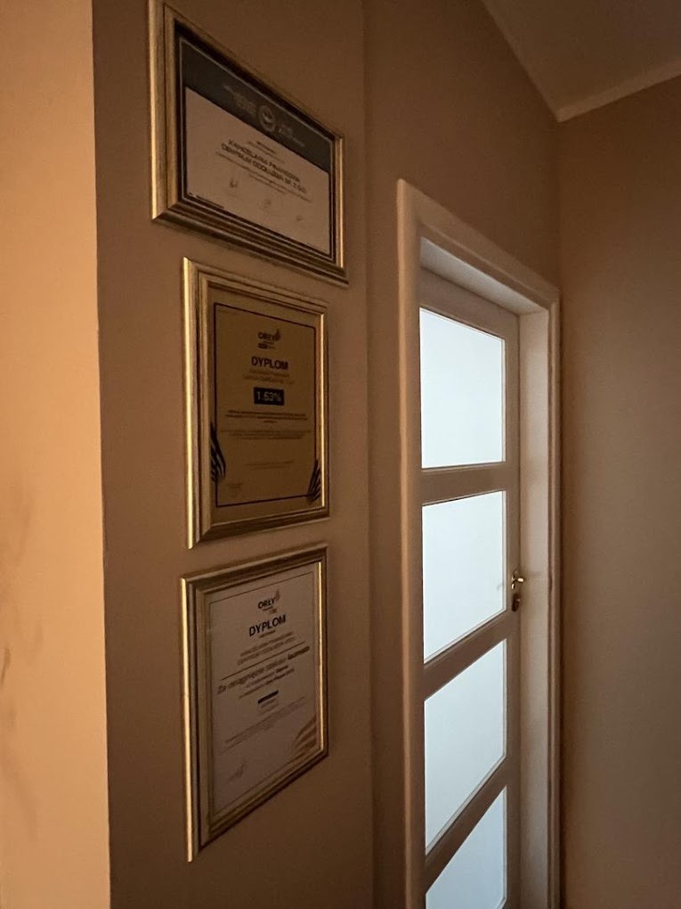 Trzy dyplomy w złotych ramach wiszą na beżowej ścianie obok białych drzwi z szybami. Dyplomy potwierdzają kompetencje.