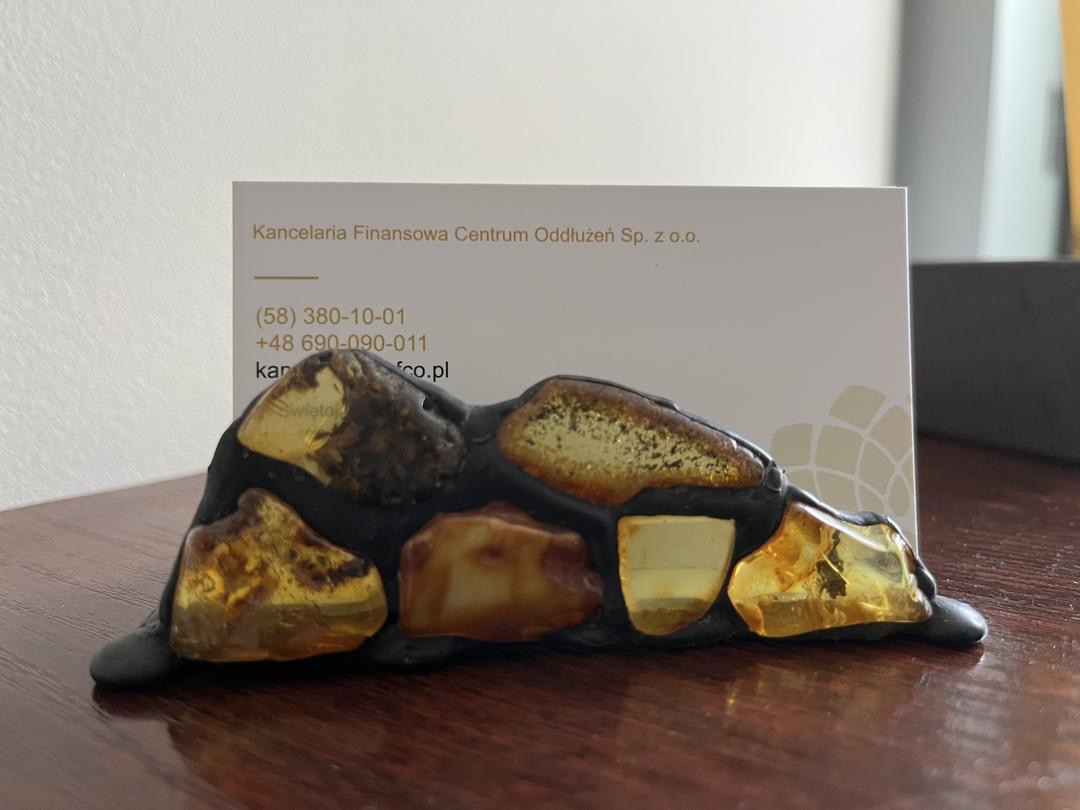 Wizytówka Kancelarii Finansowej Centrum Oddłużeń umieszczona w unikalnym, bursztynowym stojaku na drewnianym biurku. Widoczne dane kontaktowe firmy.