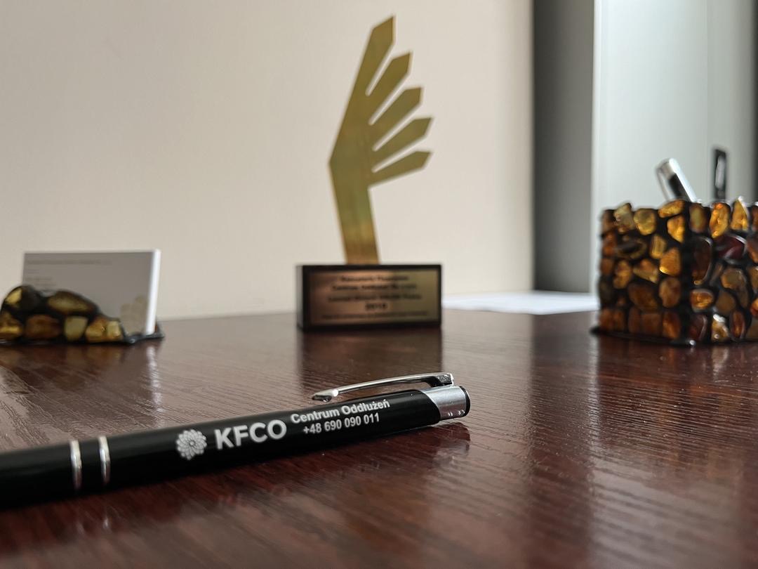 Biuro: długopis KFCO na drewnianym biurku, w tle nagroda, wizytownik z bursztynem i pojemnik na długopisy z bursztynu. Stonowana kolorystyka, blik światła na blacie.
