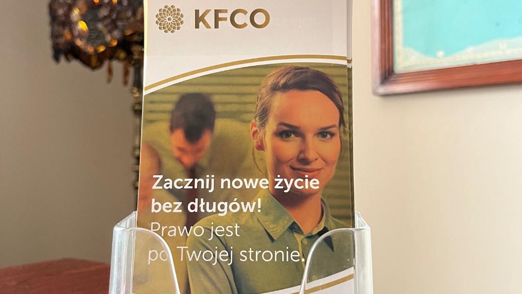 Ulotka reklamowa z hasłem 'Zacznij nowe życie bez długów! Prawo jest po Twojej stronie.' i wizerunkiem uśmiechniętej kobiety, umieszczona w przezroczystym stojaku.