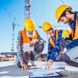 OK PERSONAL SPÓŁKA Z OGRANICZONĄ ODPOWIEDZIALNOŚCIĄ - Trzech inżynierów budowlanych w kaskach i kamizelkach analizuje plany na budowie z żurawiami w tle. Jeden z nich trzyma tablet.