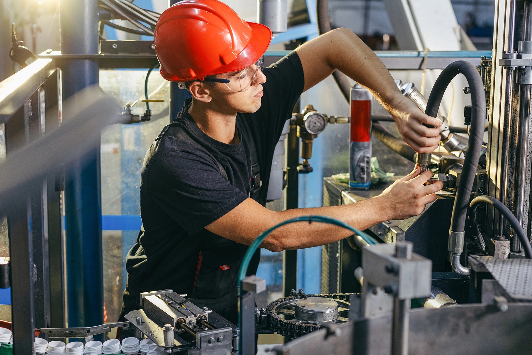Pracownik w kasku i okularach ochronnych naprawia maszynę produkcyjną. Widoczne elementy linii produkcyjnej i narzędzia. Jasne oświetlenie, skupienie na detalu.