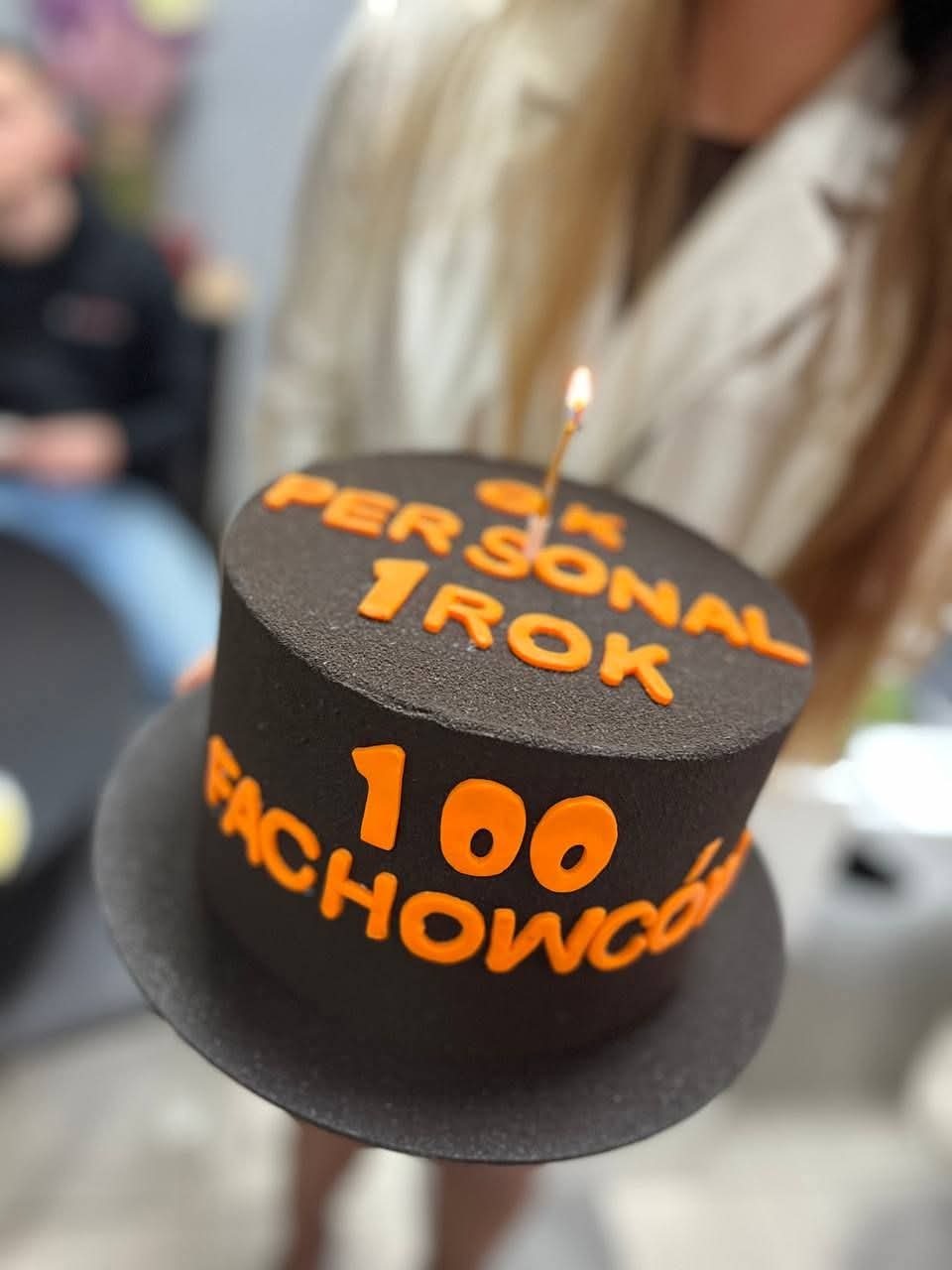 Tort w kształcie cylindra z napisem '100k Personal 1 Rok Fachowców' i płonącą świeczką, trzymany przez osobę w jasnej marynarce. Uroczystość firmowa.