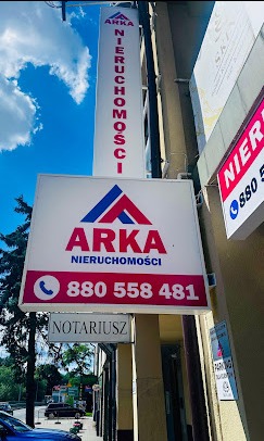 Szyld agencji nieruchomości ARKA z numerem telefonu na tle błękitnego nieba i zieleni drzew. Widoczny napis NOTARIUSZ. Ujęcie z dołu, w słoneczny dzień.