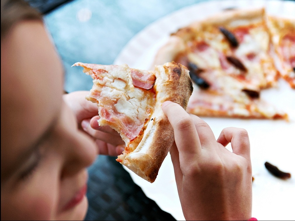 Pizza z szynką i papryczkami w dłoni dziecka, na tle talerza z resztą pizzy. Ujęcie z bliska, skupione na detalu jedzenia i interakcji.