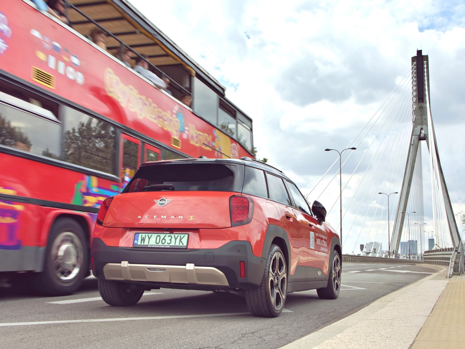 Czerwony samochód Mini Aceman S na tle mostu i autobusu turystycznego. Widok z tyłu, tablica rejestracyjna widoczna. Pochmurne niebo.