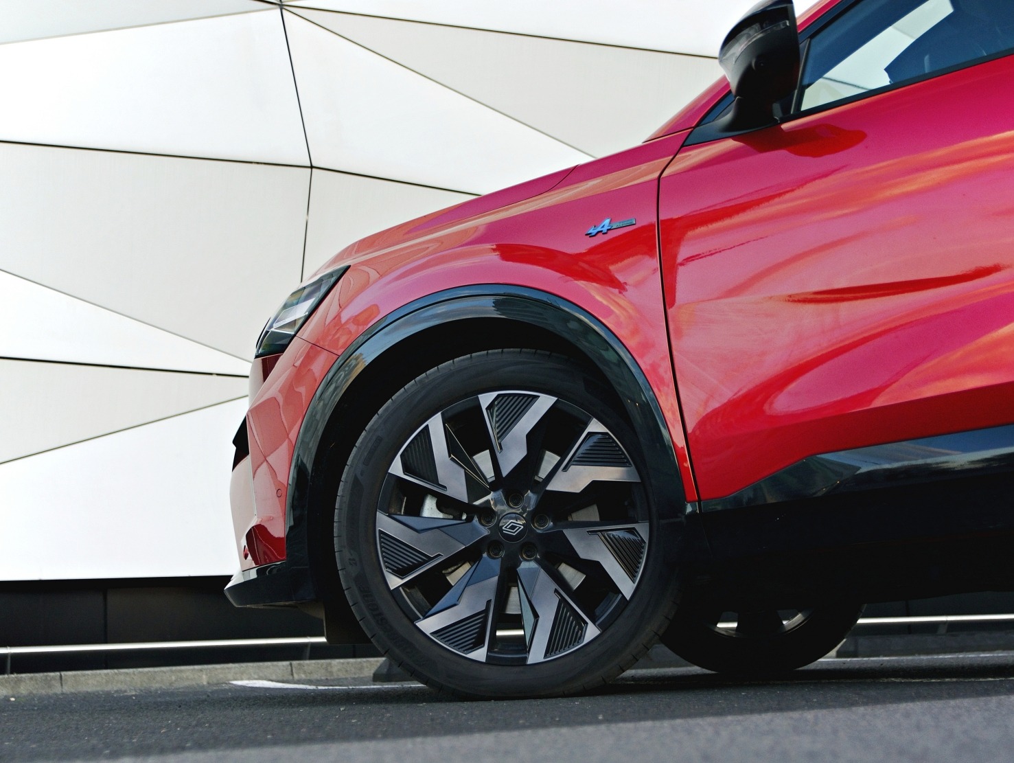 Czerwony samochód z czarnymi felgami na tle białej, geometrycznej ściany. Widoczny fragment karoserii, koło i reflektor. Ujęcie z dołu, podkreślające nowoczesny design.