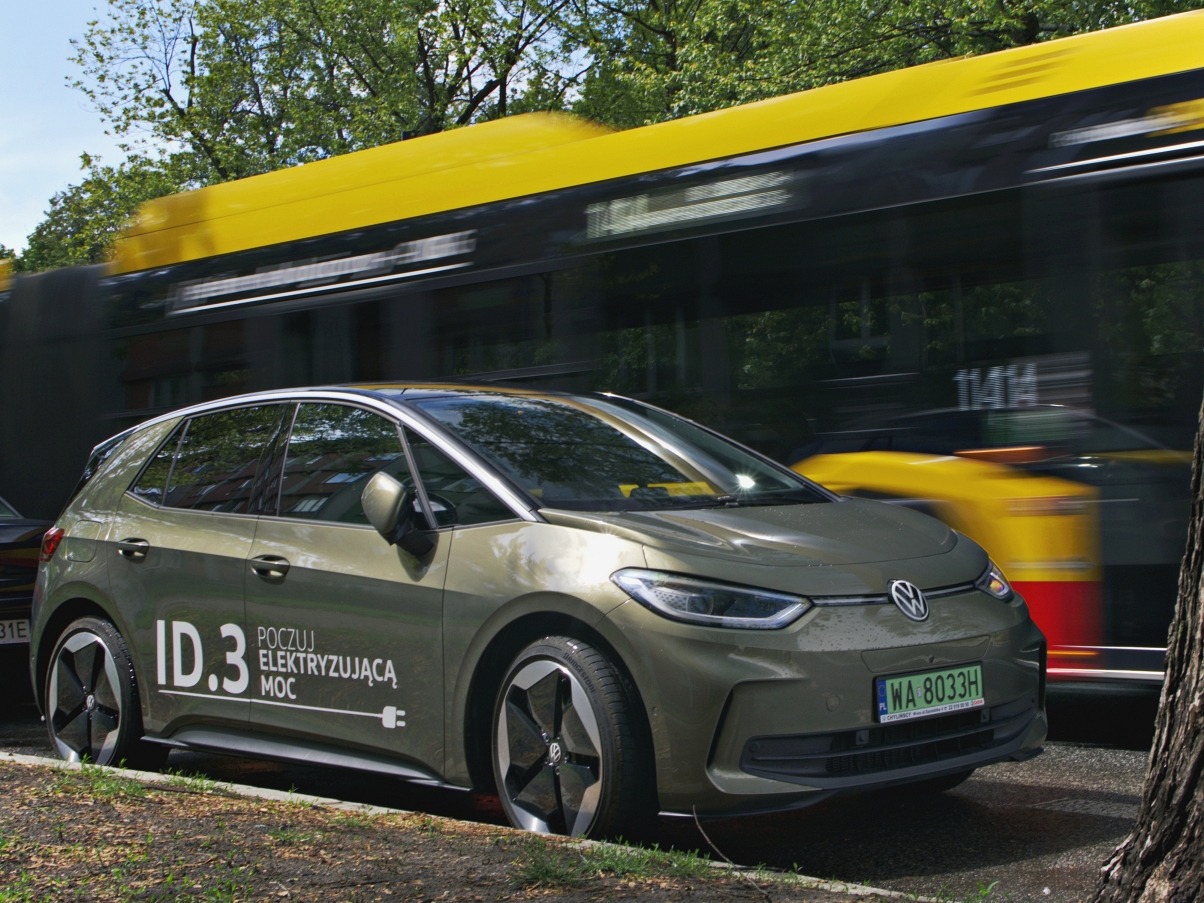Elektryczny Volkswagen ID.3 w oliwkowym kolorze na tle rozmytego, żółtego tramwaju w ruchu. Na karoserii widoczne logo i napis 'Poczuj elektryzującą moc'.