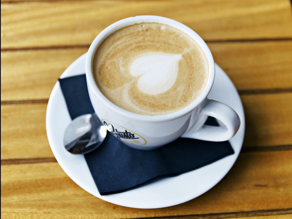 Kawa latte z sercem na piance w białej filiżance na drewnianym stole, z czarną serwetką i łyżeczką. Ujęcie z góry, ciepłe oświetlenie.