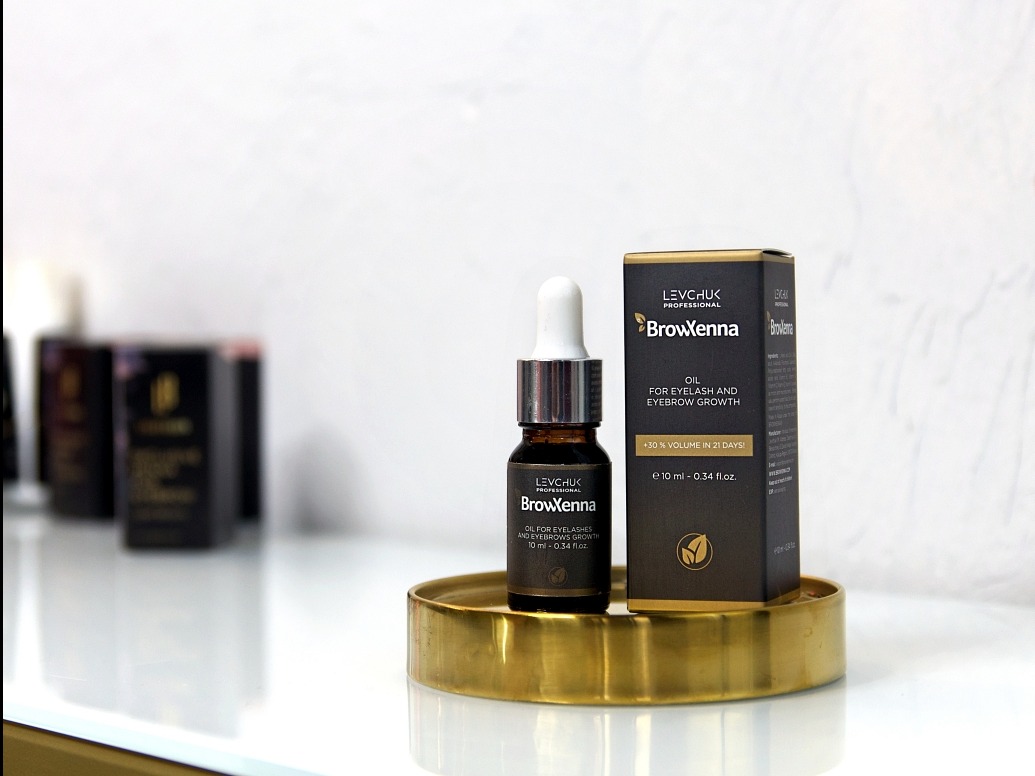 Olej BrowXenna do rzęs i brwi na złotym talerzyku, obok pudełko. Produkt do pielęgnacji z obietnicą +30% objętości w 21 dni. Stonowana kolorystyka, elegancki design.