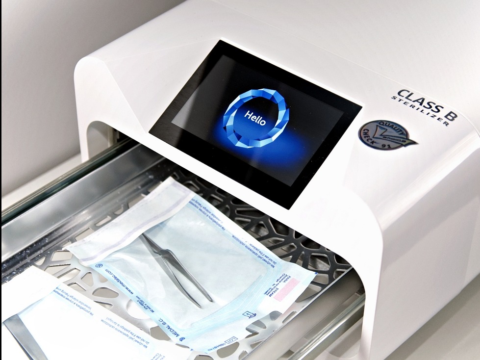 Biały autoklaw klasy B z otwartą szufladą, w której znajdują się zapakowane narzędzia. Na ekranie autoklawu widnieje napis 'Hello'.