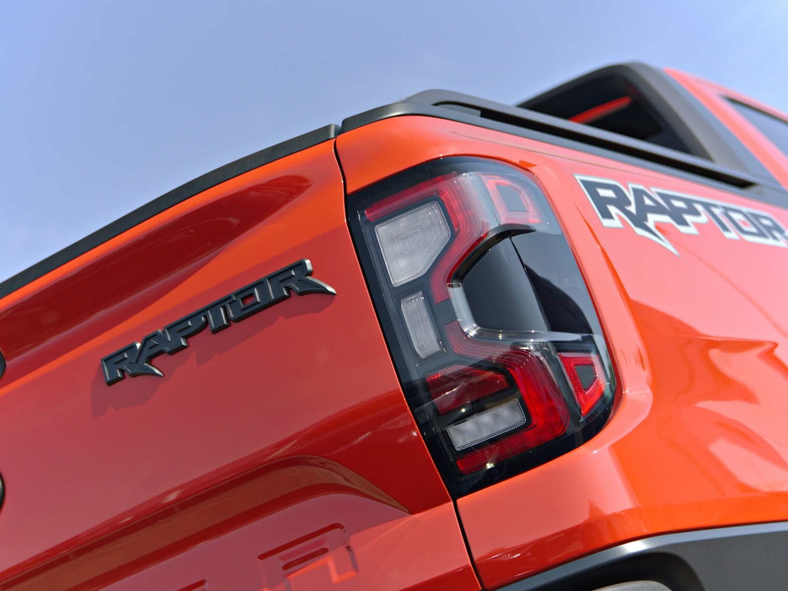 Pomarańczowy Ford Raptor z czarnym napisem 'RAPTOR' i tylną lampą, sfotografowany pod kątem w Warszawie. Widoczne detale karoserii i oświetlenia.