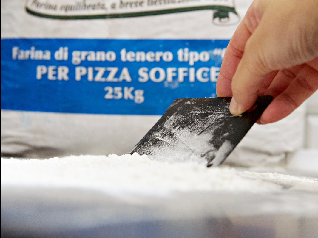 Nabieranie mąki do pizzy szpatułką z worka 25kg. Mąka typu '00' do pizzy soffice. Ujęcie z bliska, widoczna faktura mąki i dłoń.
