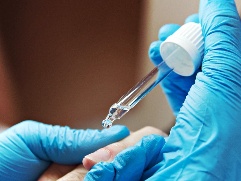 Nail stylist aplikuje serum na paznokieć klientki za pomocą pipety. Dłonie w niebieskich rękawiczkach, zbliżenie na dłoń i paznokieć, neutralne tło.