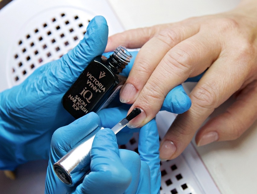 Aplikacja czarnego lakieru Victoria Vynn na paznokcie dłoni w niebieskich rękawiczkach, z lampą UV w tle. Precyzyjne malowanie blisko skórki.