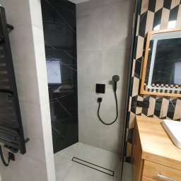 Usługi Hydrauliczne i Wykończeniowe Krzysztof Asztemborski - Nowoczesna kabina prysznicowa z czarnymi akcentami: deszczownica, panel z efektem marmuru, odpływ liniowy. Umywalka z drewnianą szafką i geometryczna ściana.