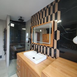 Usługi Hydrauliczne i Wykończeniowe Krzysztof Asztemborski - Nowoczesna łazienka z drewnianą szafką pod umywalką, lustrem z podświetleniem i kabiną prysznicową typu walk-in. Na ścianie dekoracyjne płytki w geometryczny wzór.