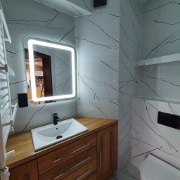 Usługi Hydrauliczne i Wykończeniowe Krzysztof Asztemborski - Nowoczesna łazienka z płytkami imitującymi marmur, drewnianą szafką pod umywalką i podświetlanym lustrem. Widoczny grzejnik drabinkowy i toaleta.