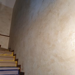 Usługi Hydrauliczne i Wykończeniowe Krzysztof Asztemborski - Beżowa ściana z dekoracyjną fakturą w korytarzu schodowym. Schody zabezpieczone folią malarską. Widoczny fragment drewnianej listwy przypodłogowej.