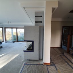 Usługi Hydrauliczne i Wykończeniowe Krzysztof Asztemborski - Wnętrze w trakcie prac: obudowa kominka z płyt karton-gips, zabezpieczona podłoga, duże okna z widokiem na zewnątrz. Widoczny etap budowy.