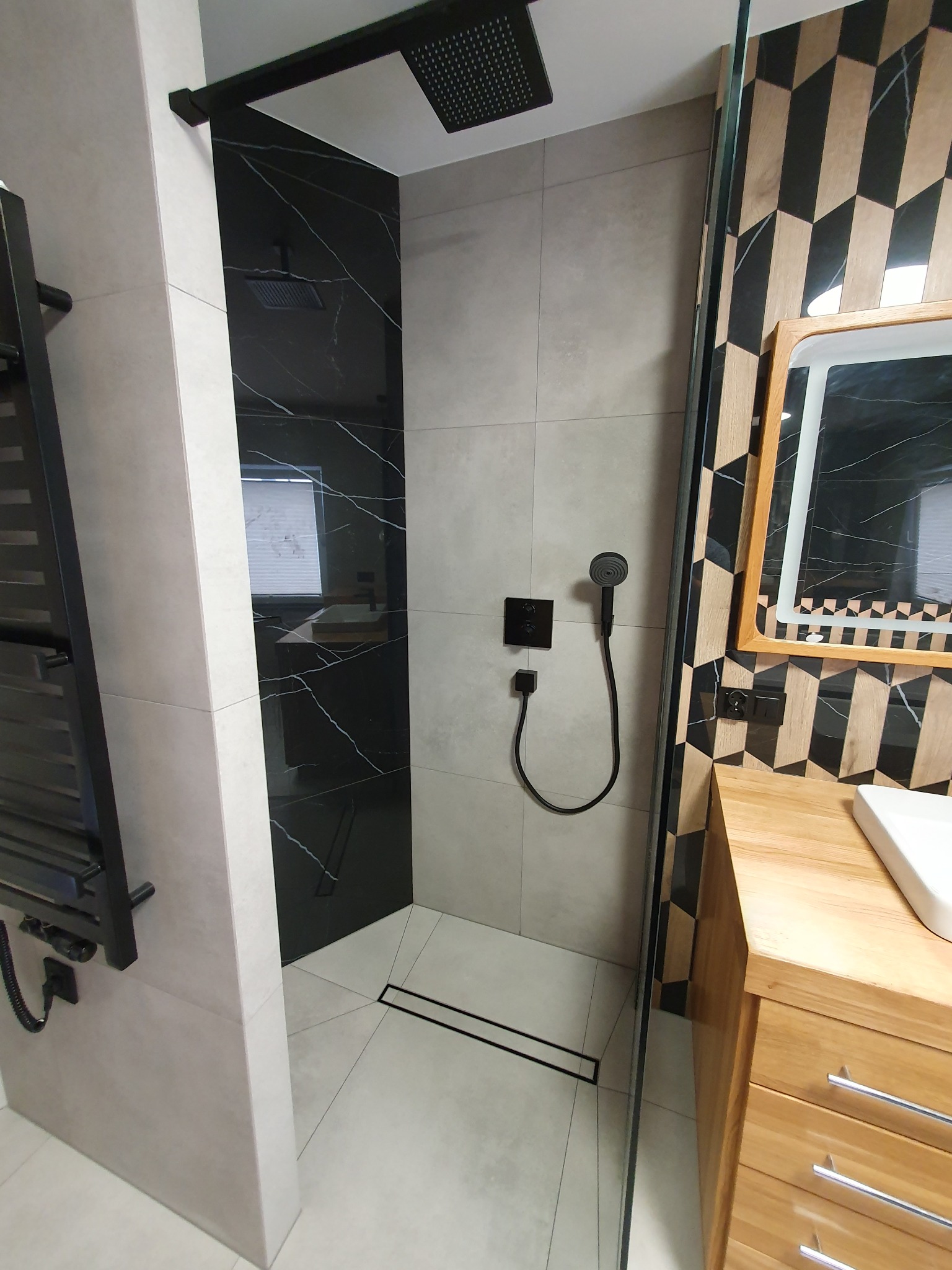Nowoczesna kabina prysznicowa z czarnymi akcentami: deszczownica, panel z efektem marmuru, odpływ liniowy. Umywalka z drewnianą szafką i geometryczna ściana.