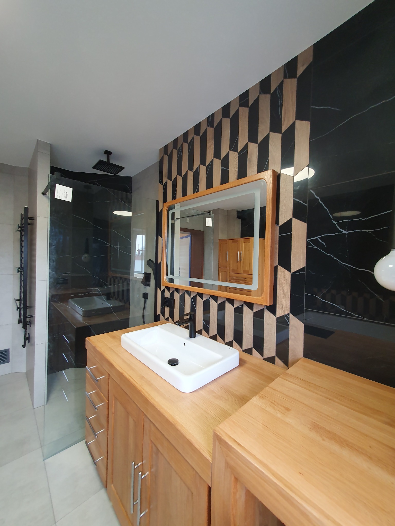 Nowoczesna łazienka z drewnianą szafką pod umywalką, lustrem z podświetleniem i kabiną prysznicową typu walk-in. Na ścianie dekoracyjne płytki w geometryczny wzór.