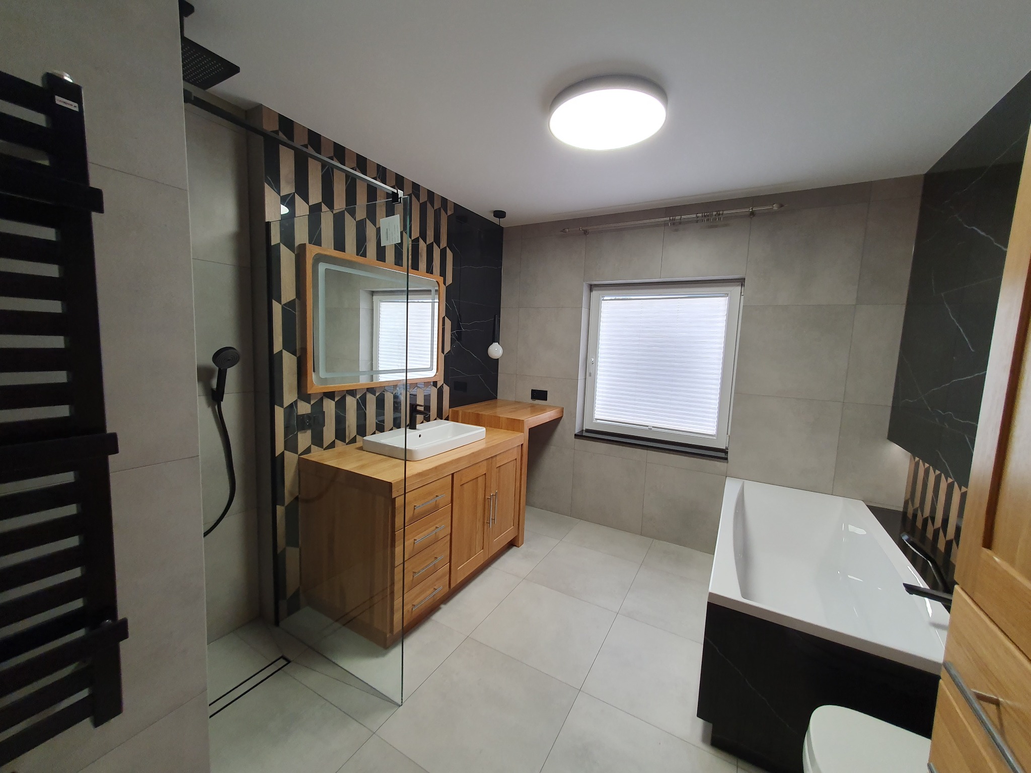 Nowoczesna łazienka z drewnianą szafką pod umywalką, kabiną prysznicową typu walk-in i wanną obudowaną czarnym marmurem. Geometryczny wzór na ścianie dodaje charakteru.