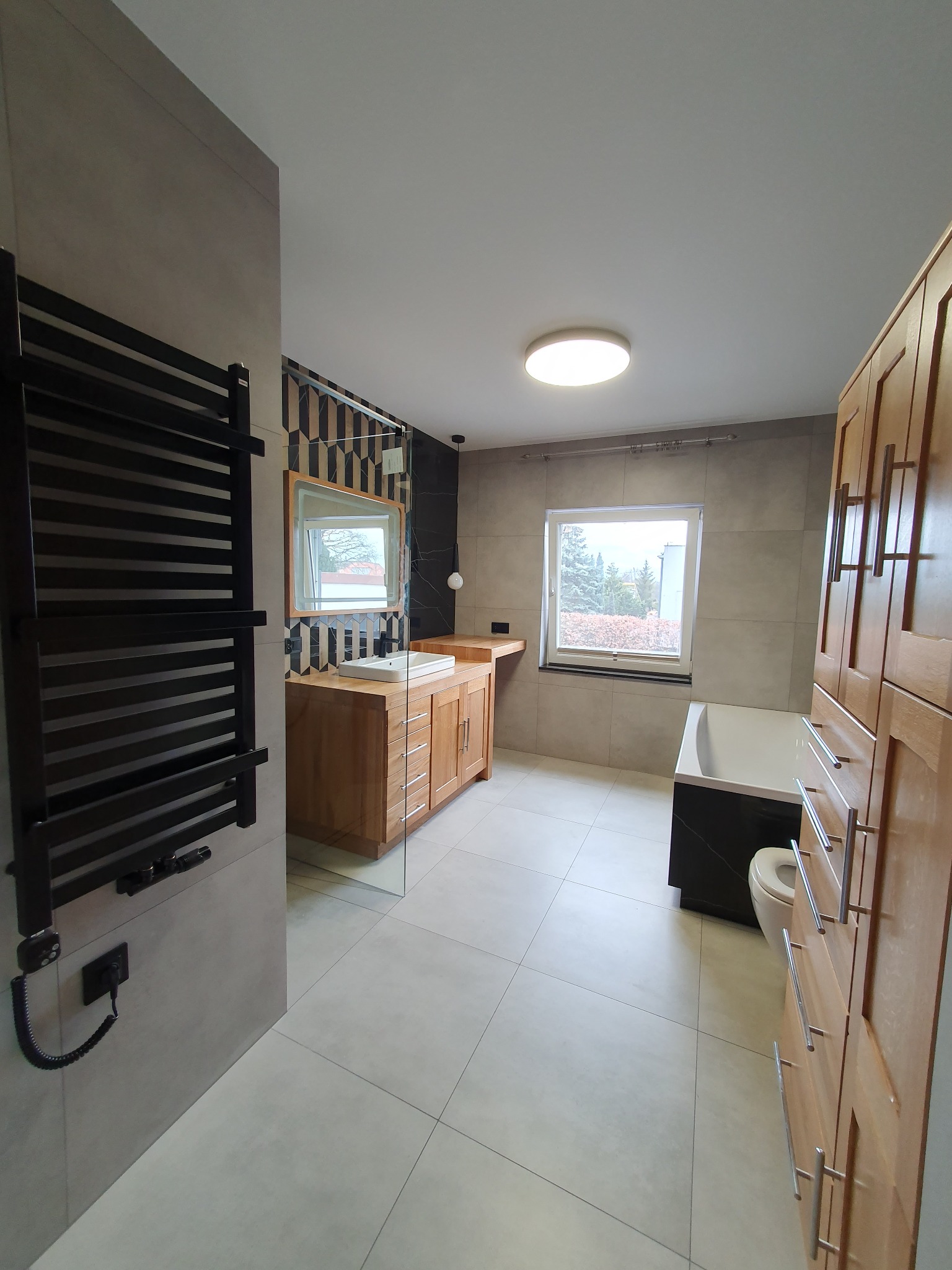 Nowoczesna łazienka z drewnianą szafką pod umywalką, grzejnikiem drabinkowym i płytkami w odcieniach szarości. Widoczne okno z widokiem na zewnątrz.