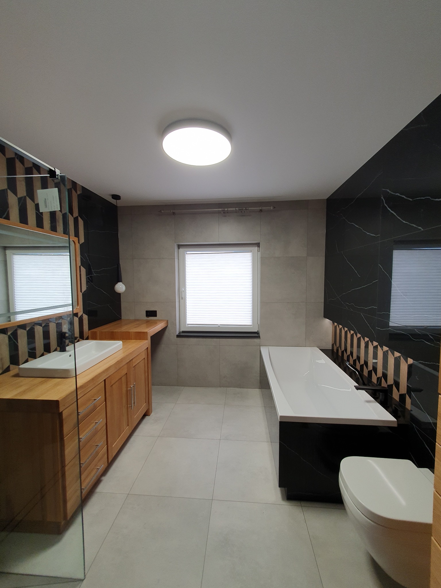 Nowoczesna łazienka z drewnianą szafką pod umywalką, czarnymi płytkami imitującymi marmur i białą wanną. Widoczne geometryczne wzory i subtelne oświetlenie.