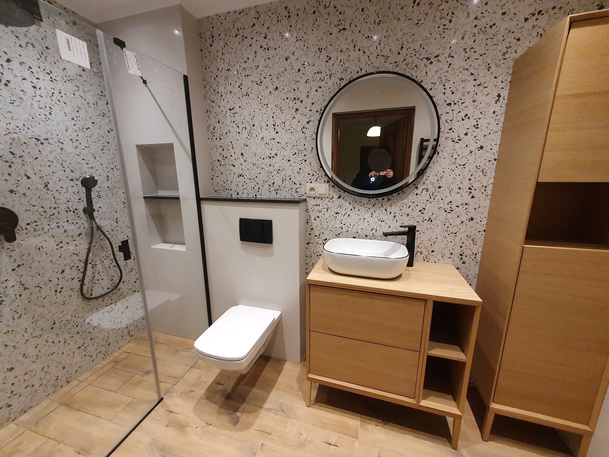 Nowoczesna łazienka z kabiną prysznicową, umywalką na drewnianej szafce i lustrem w okrągłej ramie. Ściany wykończone płytkami imitującymi lastryko.