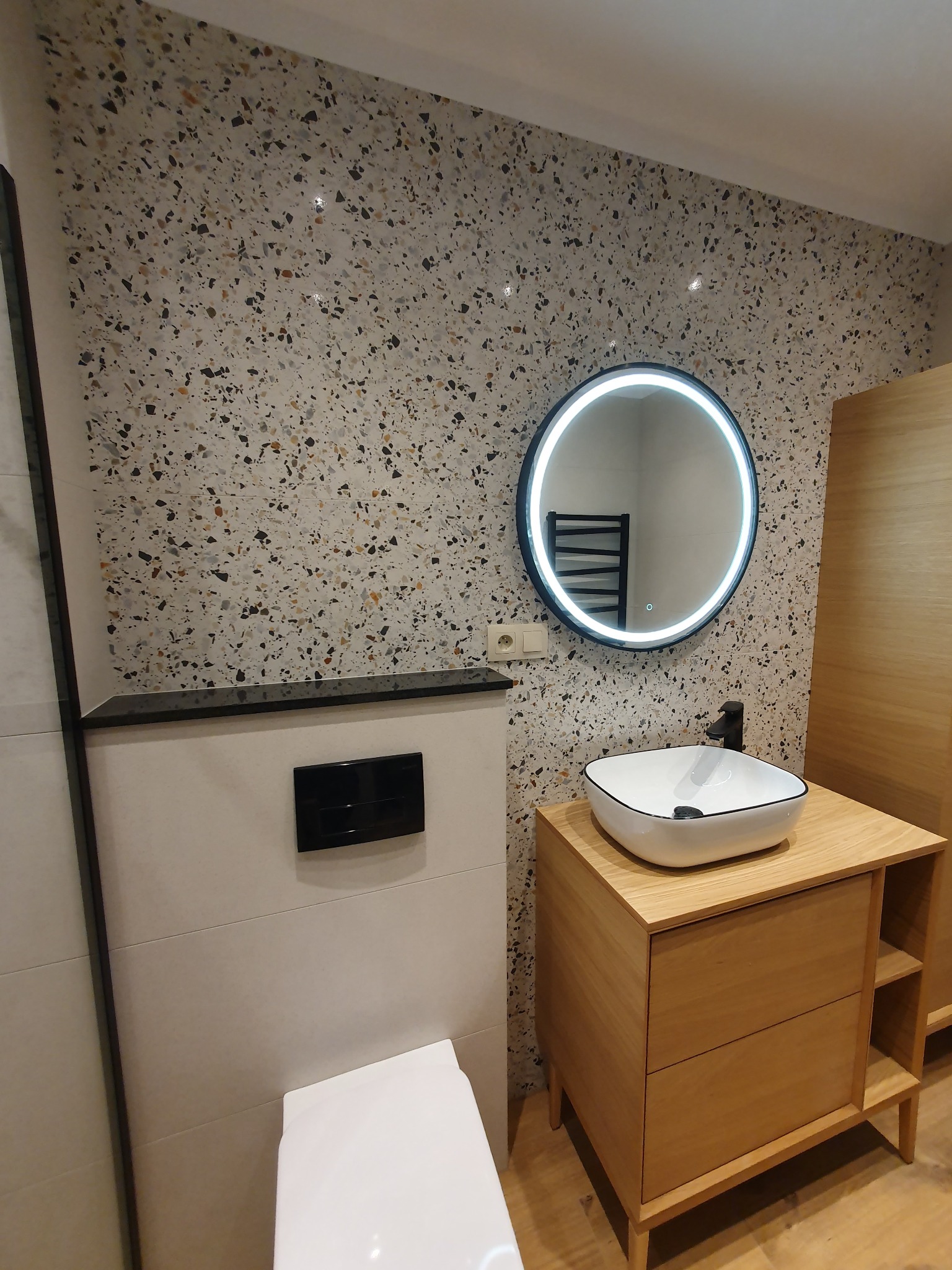 Nowoczesna łazienka z umywalką na drewnianej szafce, okrągłym lustrem z podświetleniem LED i ścianą wykończoną płytkami imitującymi lastryko.