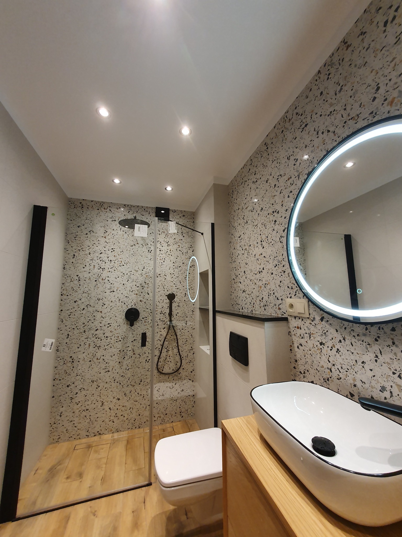 Nowoczesna łazienka z kabiną prysznicową typu walk-in, umywalką nablatową i okrągłym lustrem z podświetleniem LED. Ściany wykończone płytkami imitującymi lastryko.