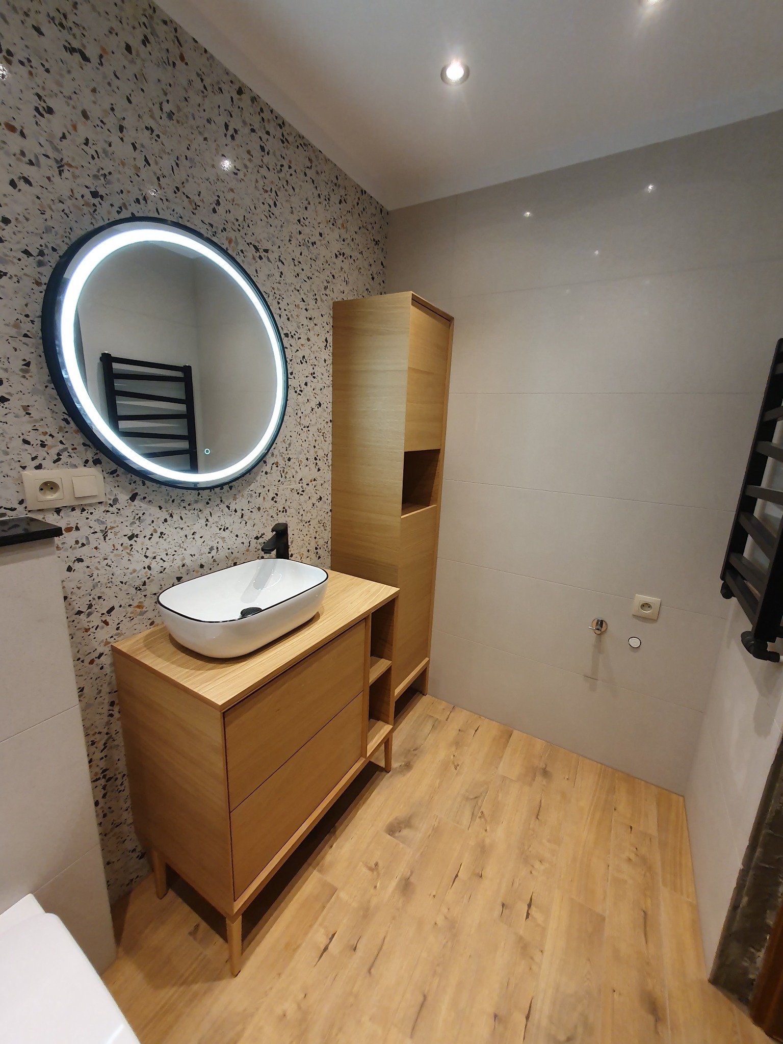 Nowoczesna łazienka z umywalką nablatową, okrągłym lustrem z podświetleniem LED i drewnianymi szafkami. Ściana z mozaiką i płytki imitujące drewno na podłodze.