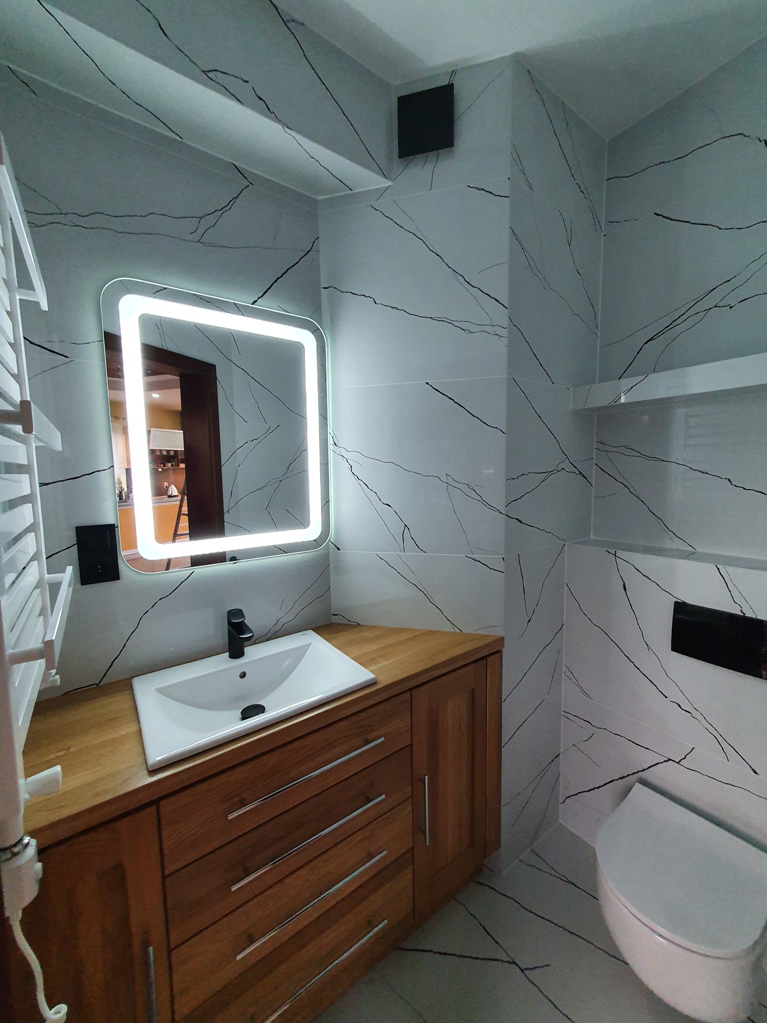Nowoczesna łazienka z płytkami imitującymi marmur, drewnianą szafką pod umywalką i podświetlanym lustrem. Widoczny grzejnik drabinkowy i toaleta.