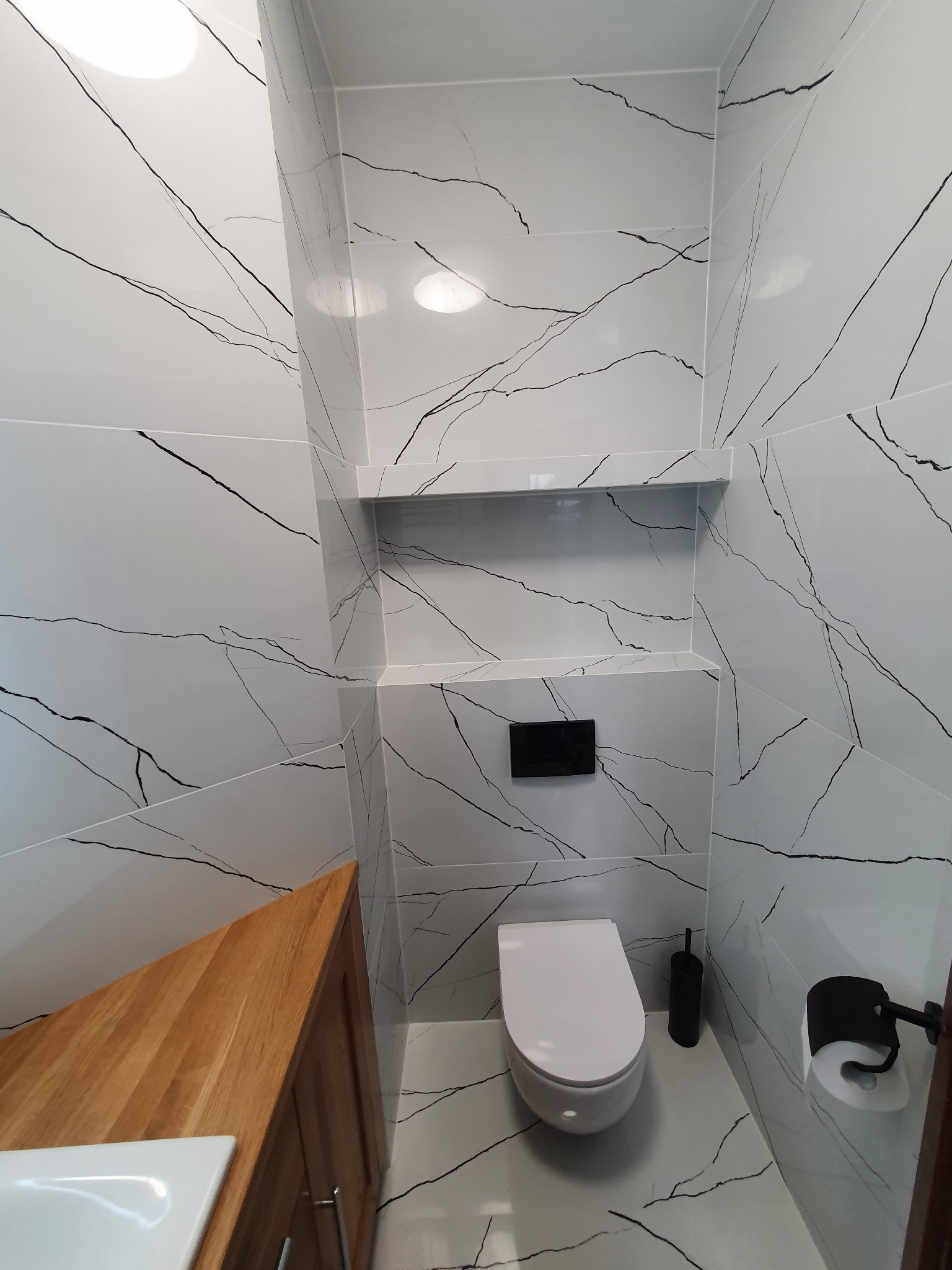 Nowoczesna łazienka z białymi płytkami imitującymi marmur, podwieszana toaleta, drewniana szafka pod umywalką i wnęka w ścianie. Minimalistyczny design.