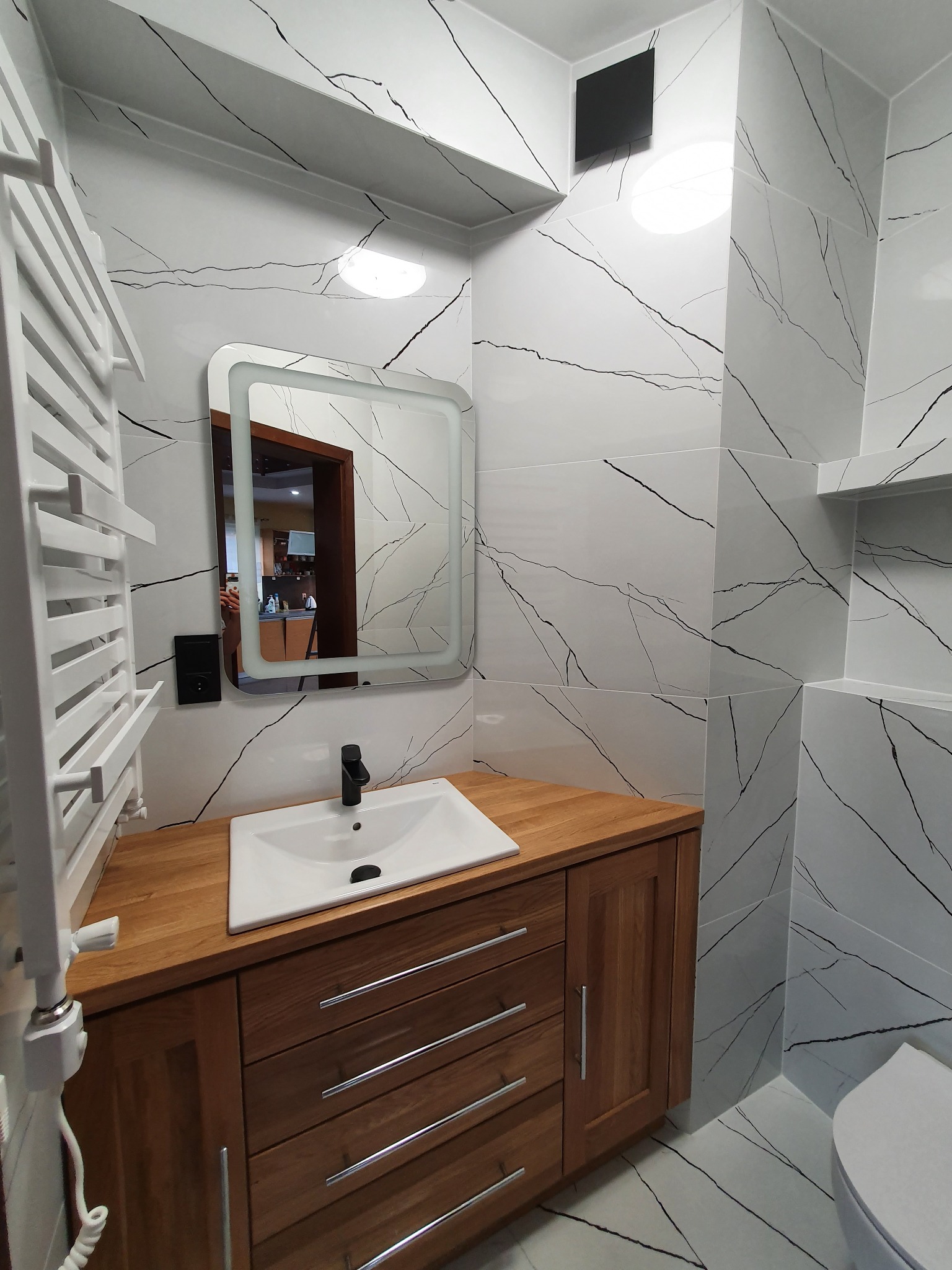 Nowoczesna łazienka z drewnianą szafką pod umywalką, białymi płytkami imitującymi marmur i podświetlanym lustrem. Widoczny grzejnik drabinkowy i fragment toalety.