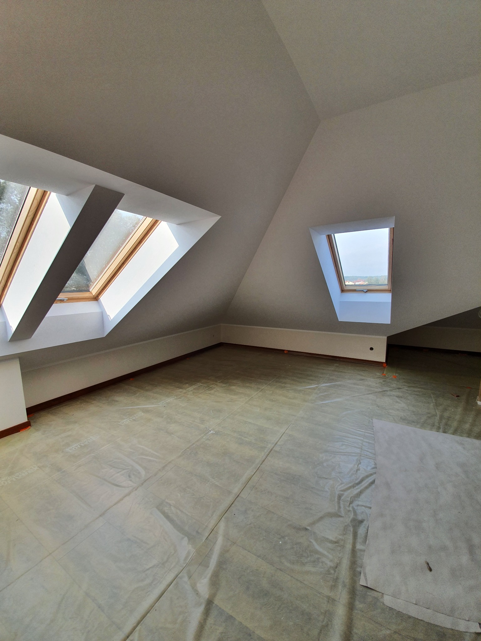 Poddasze ze skosami i oknami dachowymi, przygotowane do prac wykończeniowych. Podłoga zabezpieczona folią ochronną. Ściany i sufit pomalowane na biało.