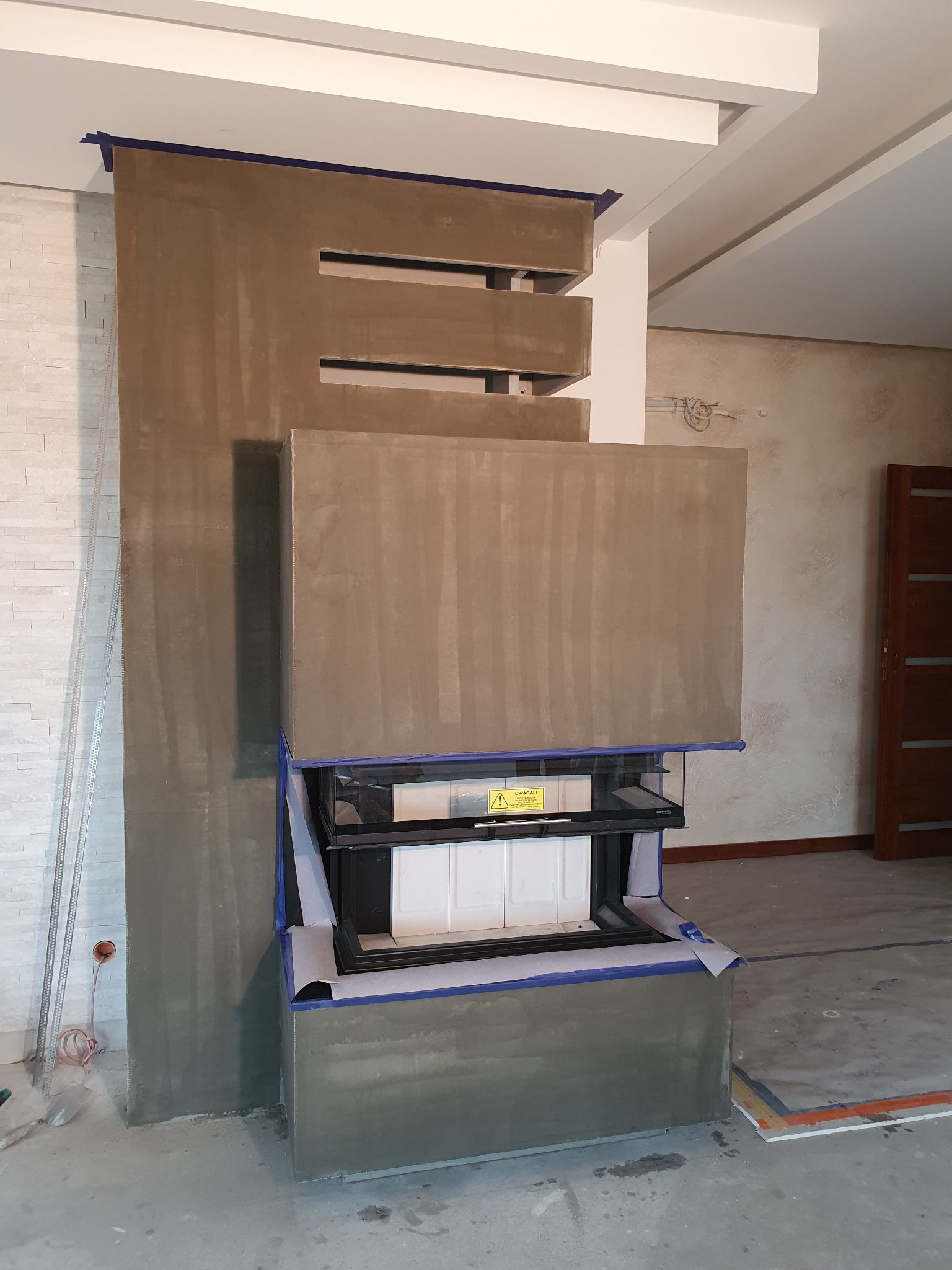 Wnętrze z kominkiem w trakcie obudowy, zabezpieczonym taśmą malarską. Widoczne surowe wykończenie ściany i elementy konstrukcyjne. W tle ściana z dekoracyjną fakturą.