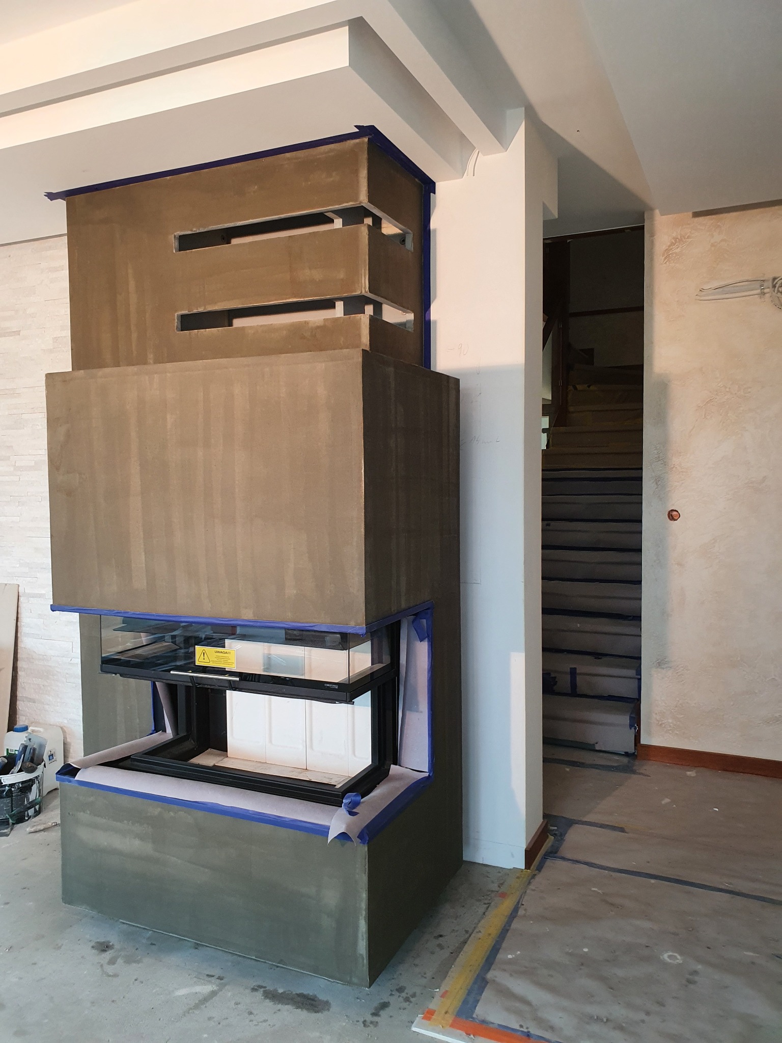 Obudowa kominka w trakcie prac wykończeniowych, zabezpieczona taśmą malarską. Widoczne surowe wykończenie i fragment schodów w tle.