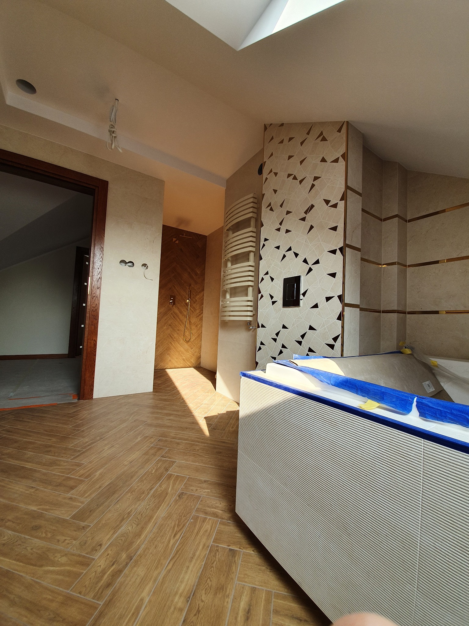 Ukończona łazienka na poddaszu z drewnopodobną podłogą w jodełkę, beżowymi płytkami i dekoracyjną ścianą z motywem trójkątów. Widoczny grzejnik i fragment wanny.