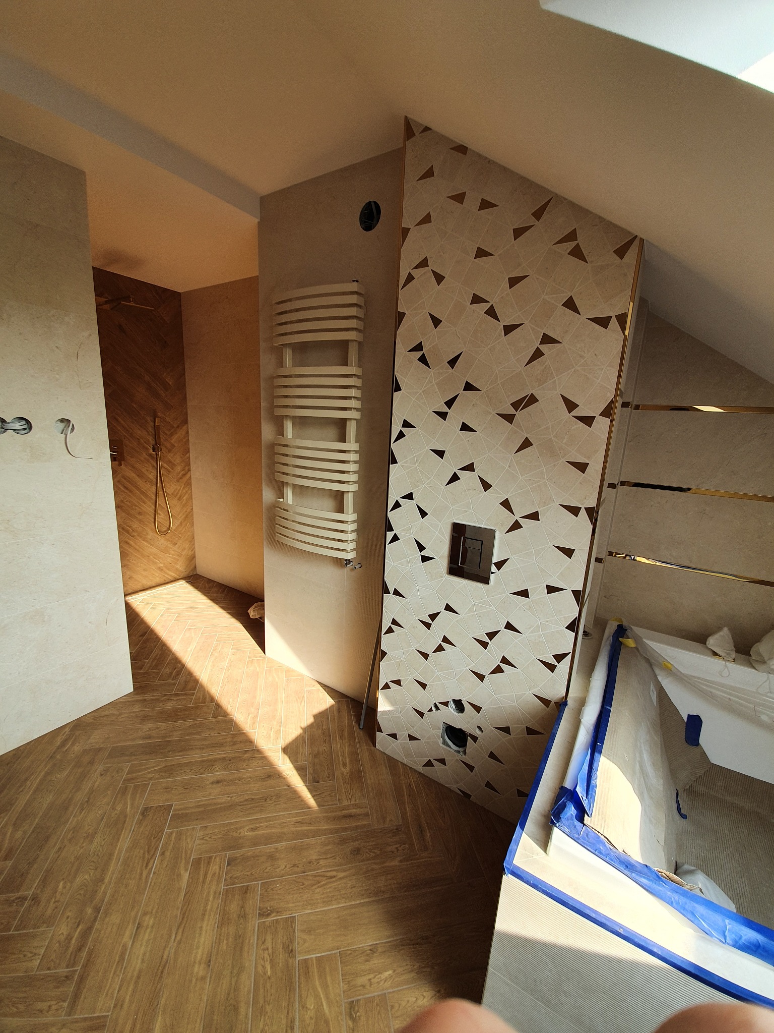 Nowoczesna łazienka w beżach: drewnopodobna podłoga w jodełkę, dekoracyjna ściana z trójkątami i grzejnik drabinkowy. Widoczny fragment wanny zabezpieczonej folią.