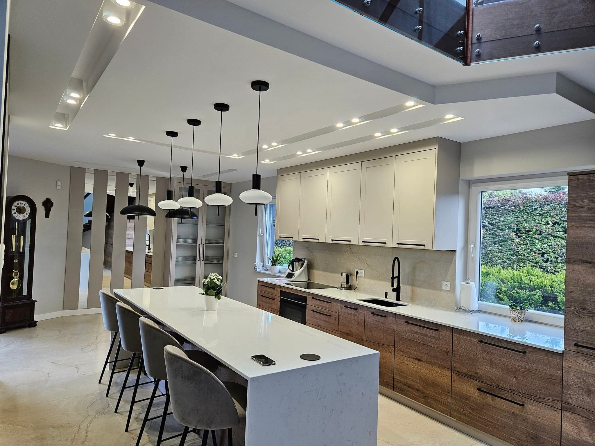 Nowoczesna kuchnia z wyspą, drewnianymi szafkami i białym blatem. Nad wyspą wiszą czarne lampy. Widoczne okno z zielenią i zegar stojący. Całość w jasnych barwach.