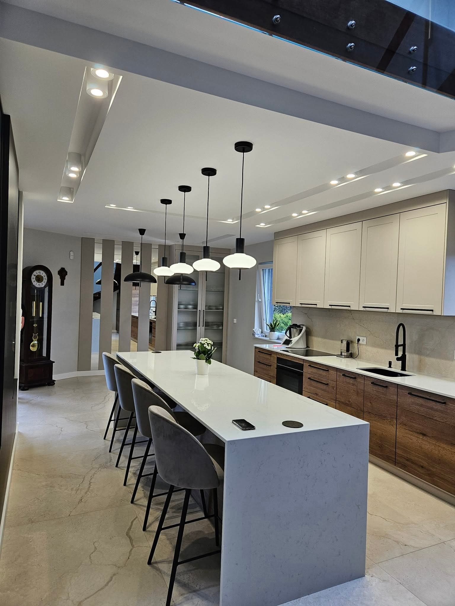 Nowoczesna kuchnia z wyspą, szarymi krzesłami barowymi i oświetleniem punktowym. Widoczny zegar stojący i przeszklona ściana. Użyto naturalnych materiałów.