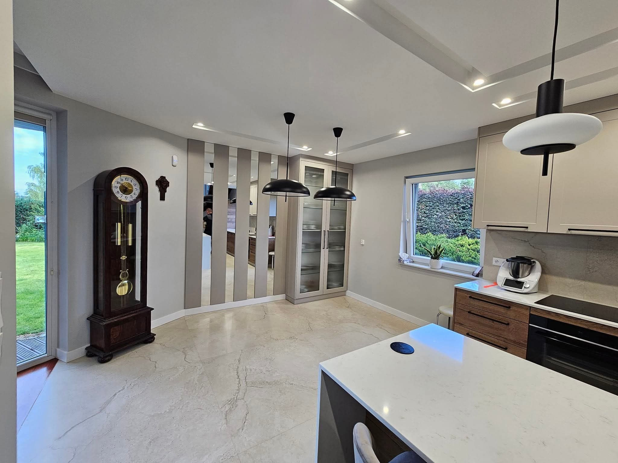Elegancka kuchnia z marmurową podłogą, wyspą i zegarem stojącym. Lustrzane panele dodają przestrzeni. Nowoczesne oświetlenie i sprzęt AGD.