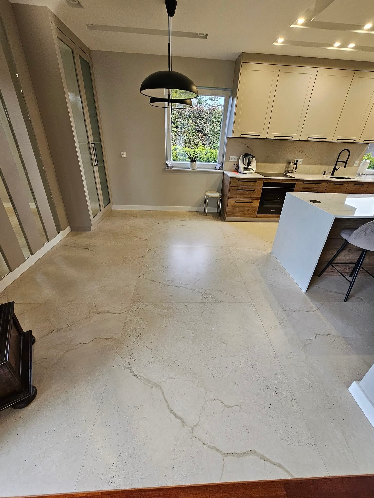 Jasne, marmurkowe płytki podłogowe w przestronnej kuchni z beżowymi szafkami i wyspą. Widok na okno z zielenią i nowoczesne oświetlenie.