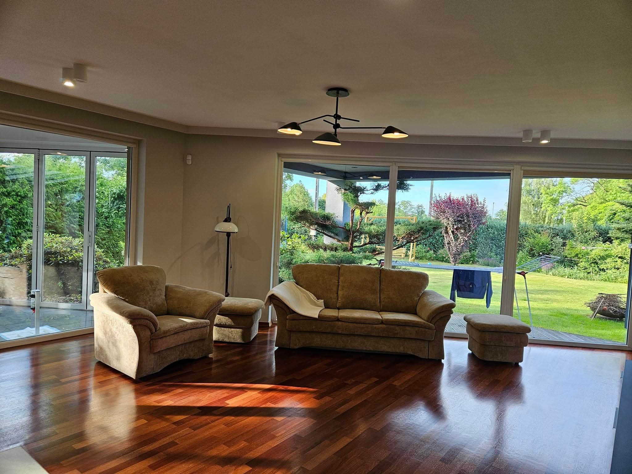 Salon z parkietem w jodełkę, beżową sofą i fotelem. Duże okna z widokiem na ogród. Nowoczesna lampa sufitowa i punktowe oświetlenie. Jasne, przestronne wnętrze.