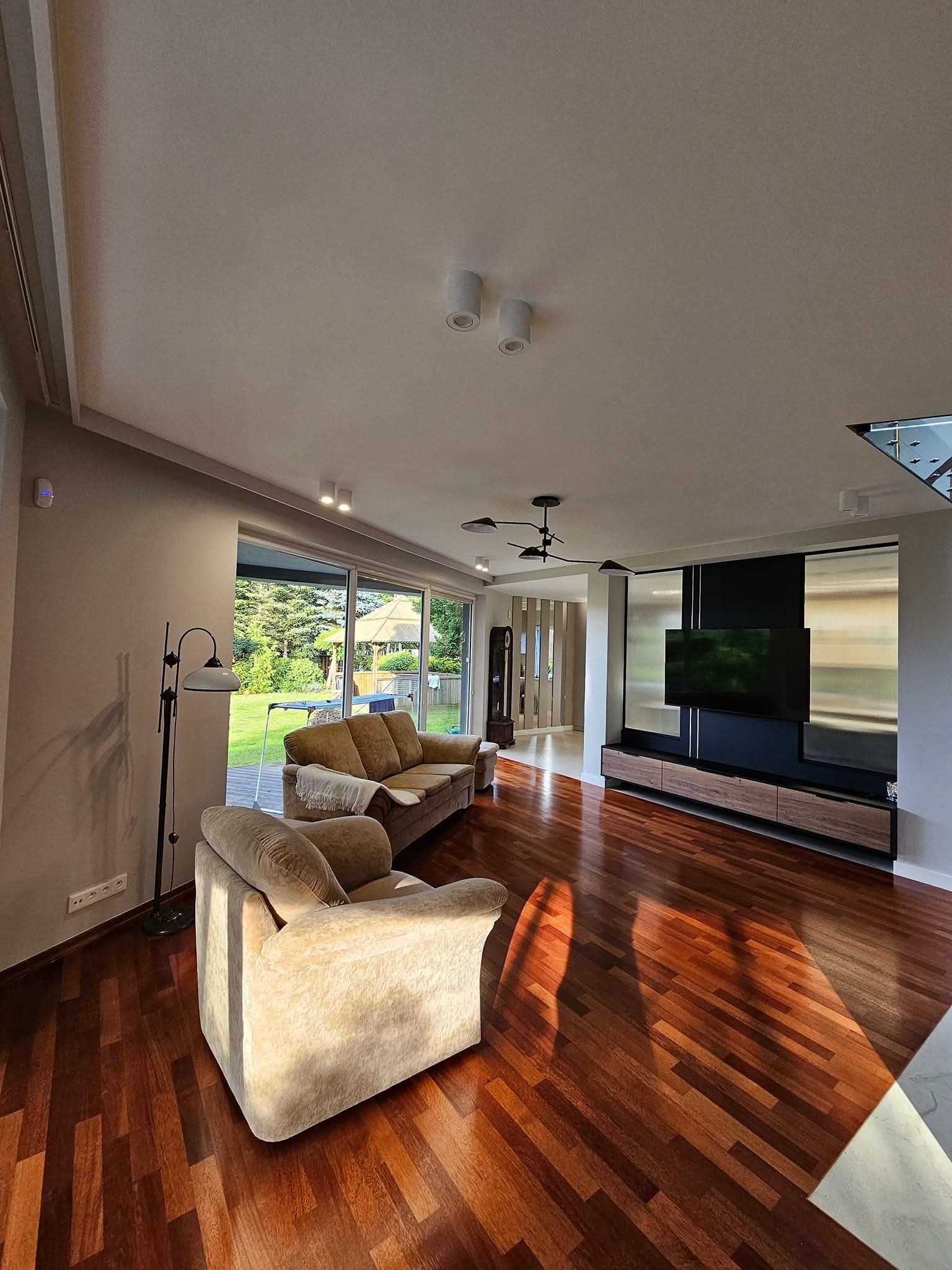 Jasne wnętrze salonu z drewnianą podłogą, beżowymi meblami i dużym oknem z widokiem na ogród. Nowoczesna aranżacja z elementami dekoracyjnymi i oświetleniem.